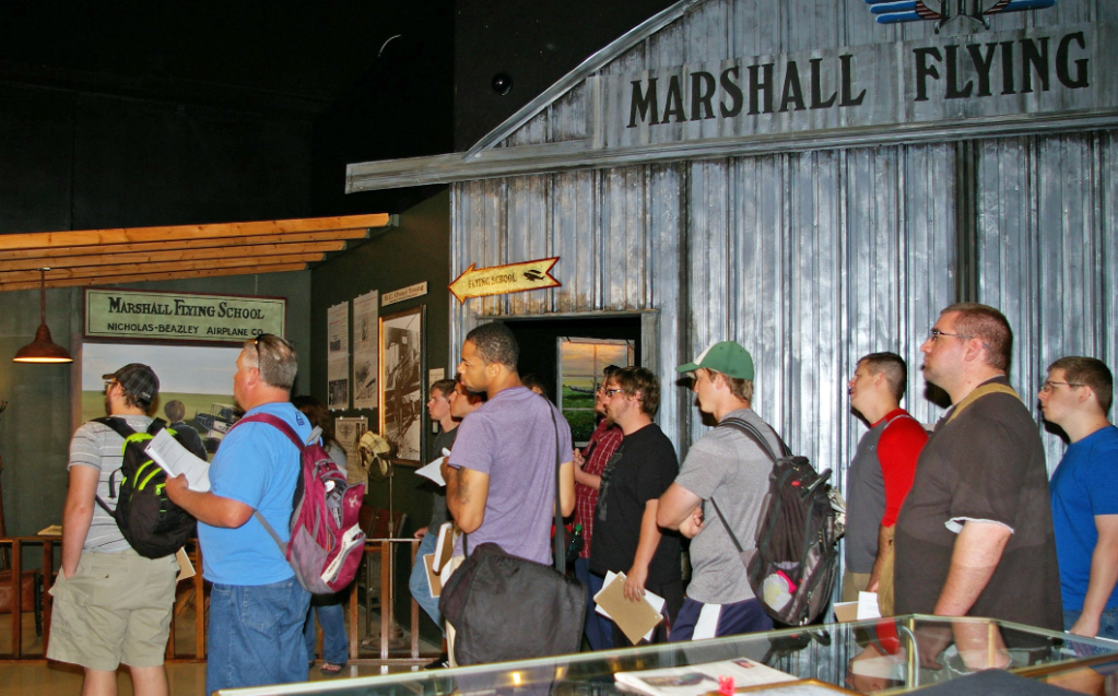 Image of a dozen students participating in a museum tour. They are standing in front of displays with a mock-up of an airfield hanger and a 1920s airplane engine.